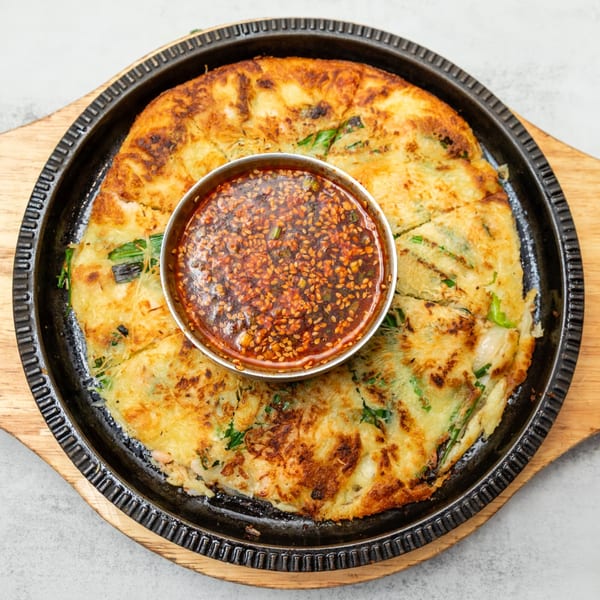 A Korean seafood pancake (haemul pajeon) with a side of dipping sauce, served on a hot plate.
